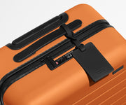 The Bigger Carry-On suitcase in Sorbet Orange shown shown from above highlighting TSA-approved combination lock and recycled leather luggage tag