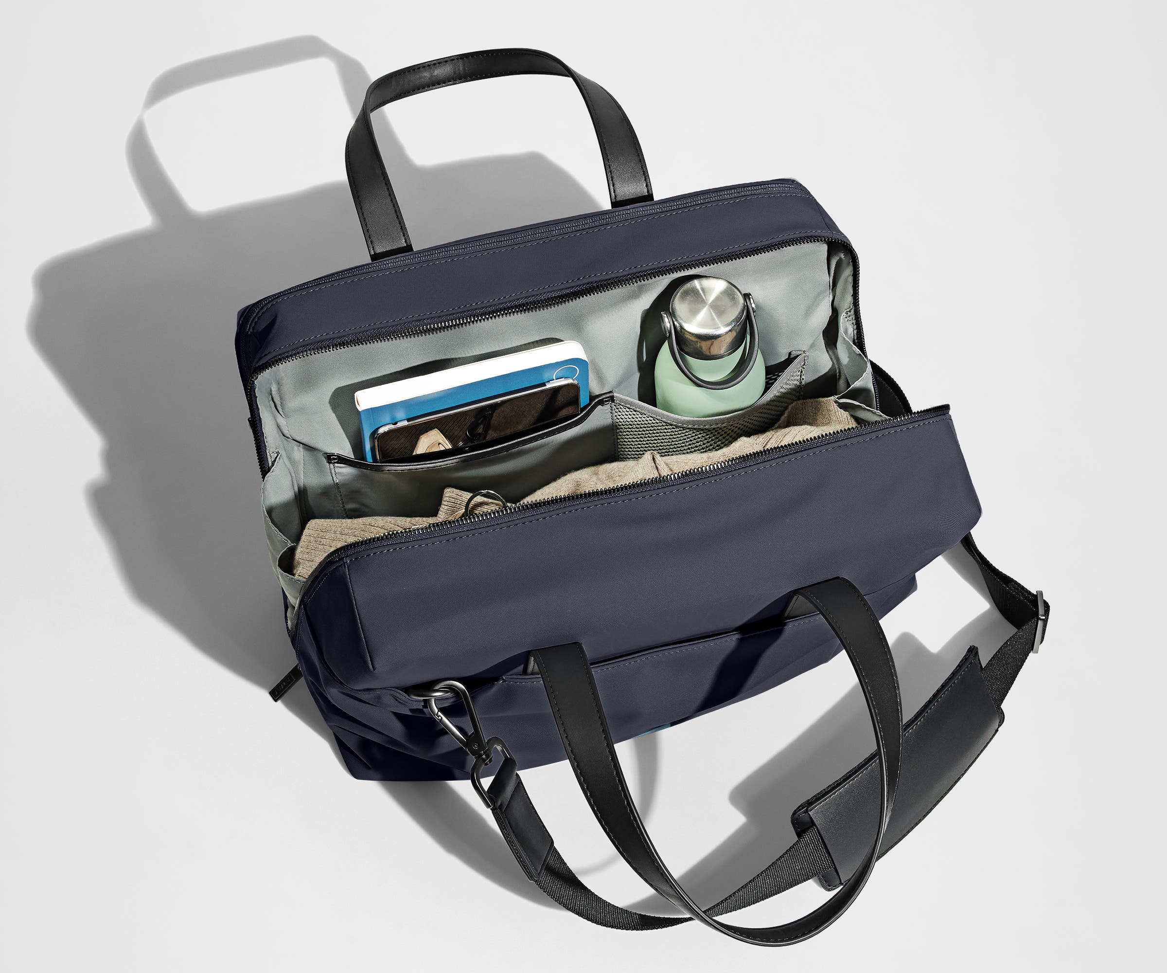 Main compartment packed with a light sweater, notebook, tablet, and water bottle. The detachable shoulder strap is clipped onto the bag.