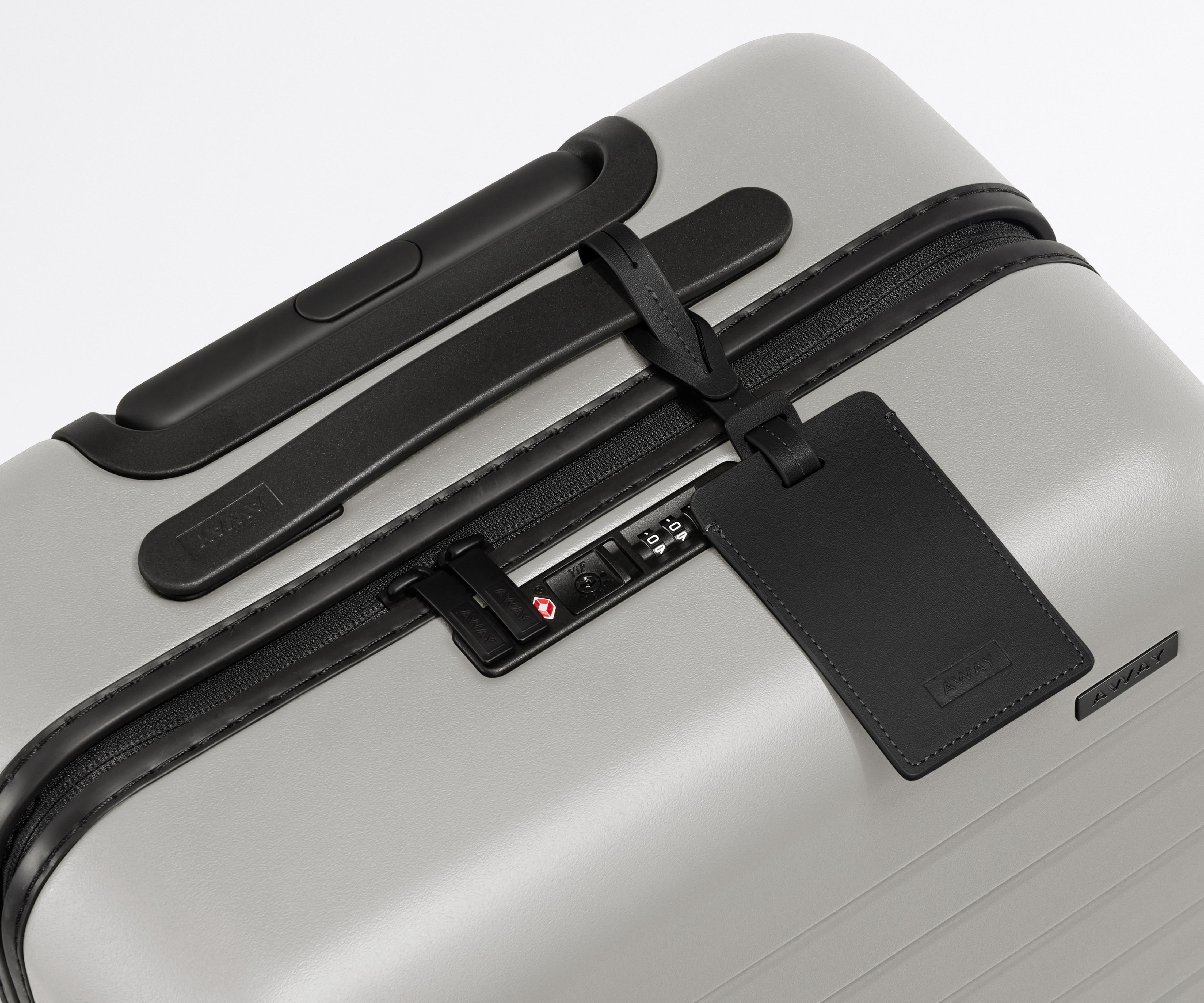 The Bigger Carry-On suitcase in Cloud Gray shown shown from above highlighting TSA-approved combination lock and recycled leather luggage tag